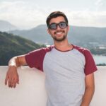 Young man in sunglasses leaning on a balcony with a scenic city view in the background.