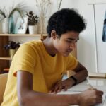 Focused African American teen studying indoors with a tablet and notebook at home.